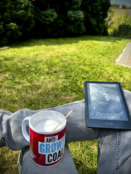 An ‘Anti-Growth Coalition’ mug and an issue of Clarkesworld magazine on a Kindle are balanced on the legs of a man fully intending to sit in his garden until all the sitting has been accomplished.