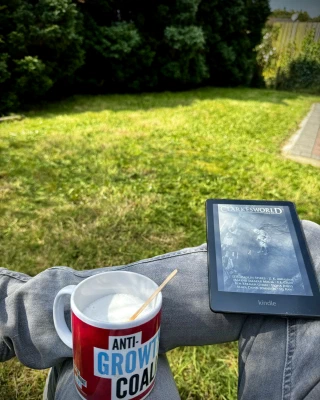 An ‘Anti-Growth Coalition’ mug and an issue of Clarkesworld magazine on a Kindle are balanced on the legs of a man fully intending to sit in his garden until all the sitting has been accomplished.