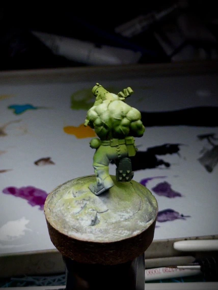 An Ork mini-onna-stick, against a background of wet palette and general painting detritus. An attempt has been made at a high-contrast, shiny, green skin. Green, yellow and purple airbrush overspray cover the rest of the surface.