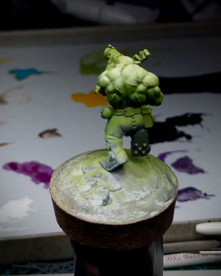 An Ork mini-onna-stick, against a background of wet palette and general painting detritus. An attempt has been made at a high-contrast, shiny, green skin. Green, yellow and purple airbrush overspray cover the rest of the surface.