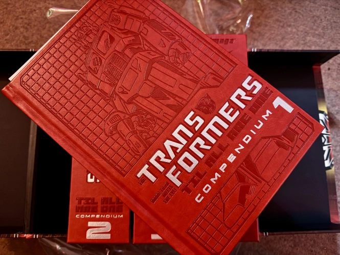 A shot showing the embossed all-red-with-white-text cover of the hardcover, volume 1 sitting on top of volumes 2-4. This is the ‘Autobot’ edition, because I am a good boy.