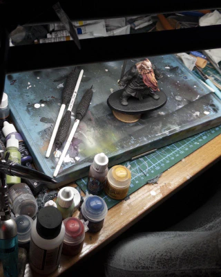 Acrylic paint pots of different sizes, oil paint tubes, paintbrushes, an airbrush and holder, putty modelling tools, bits of sprue, a covered wet palette, bits of paint-covered tissue, mugs of dirty paint water, all jumbled and crammed together as though by some passing neighbourhood madman. A mini stands in the midst, framed by a painting light and magnifying lenses at the top and a very worn faux-leather desk chair at the bottom.