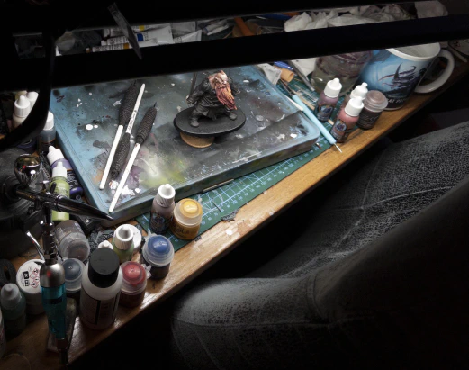 Acrylic paint pots of different sizes, oil paint tubes, paintbrushes, an airbrush and holder, putty modelling tools, bits of sprue, a covered wet palette, bits of paint-covered tissue, mugs of dirty paint water, all jumbled and crammed together as though by some passing neighbourhood madman. A mini stands in the midst, framed by a painting light and magnifying lenses at the top and a very worn faux-leather desk chair at the bottom.