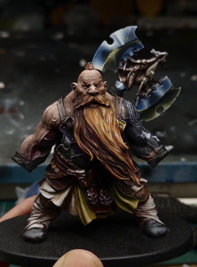 The first full body shot of Not Gotrek that we’ve seen in a while. His hair has been (temporarily) removed so that the reverse of the axe can be tacked to the wrong shoulder and viewed in comparison with the rest of the painting. Some initial reflections have been added to the tiny pieces of shiny chest armour actually visible beneath his massive beard. Eventually the two styles should hopefully match and unify the appearance a little.

Ignore the very WIP badly drawn doggo heads on his arm please. Those aren’t for you.