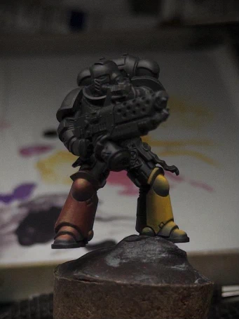 A black-primed Marine mini pictured against a wet palette of purples, yellows and pinks. The boots, shins and kneepads are coloured ‘yellow’ (stage right) and ‘red’ (stage left) in different combinations of that same paint set.