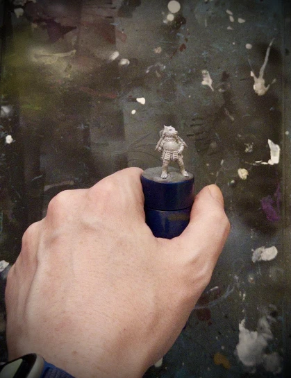 One (1) hand of approximately human proportions holding a tiny horned doggo, placed on a standard-reference painting handle. The doggo is entirely wee. It wears a cool skirt.